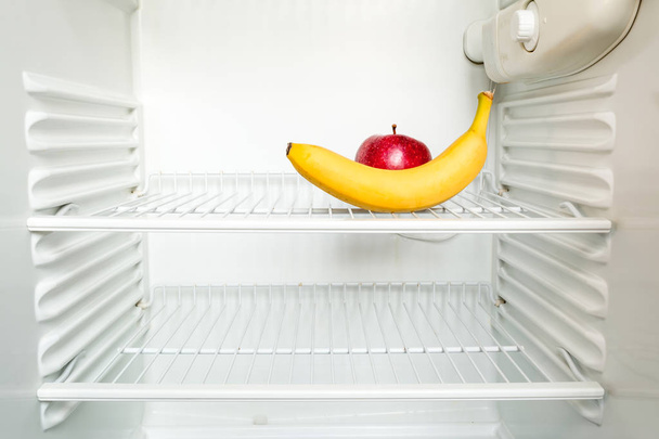 Bright fresh banana and red apple on shelf of open empty refrigerator. Weight loss diet concept. - Fotó, kép