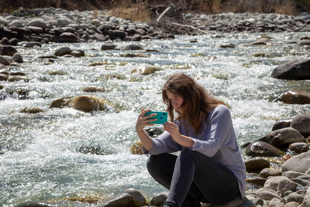 Young beautiful girl on nature takes a selfie on the phone and takes pictures of nature. Against the background of the forest and a beautiful mountain river. Tourism and travel. - Фото, зображення