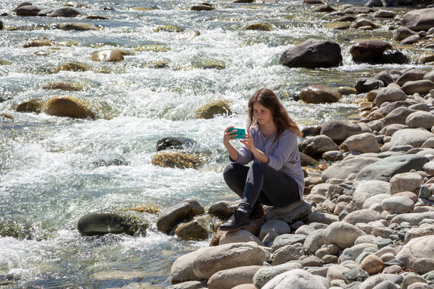 若い美少女が携帯電話で自撮りし、自然の写真を撮る。森と美しい山の川の背景に。旅行・観光. - 写真・画像