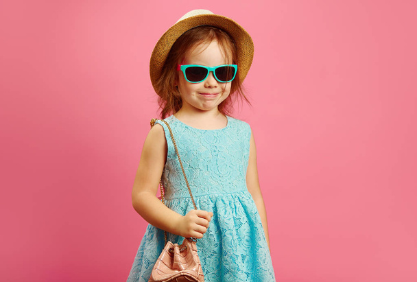 Cheerful little girl in beach clothes and handbag, stands over pink isolated background, expresses joy and happiness, dressed in beach hat and beautiful blue dress, wears sunglasses. - Foto, afbeelding