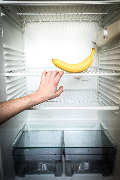 A man's hand reaches out for a single banana in an open, empty refrigerator. Weight loss diet concept. - Фото, изображение