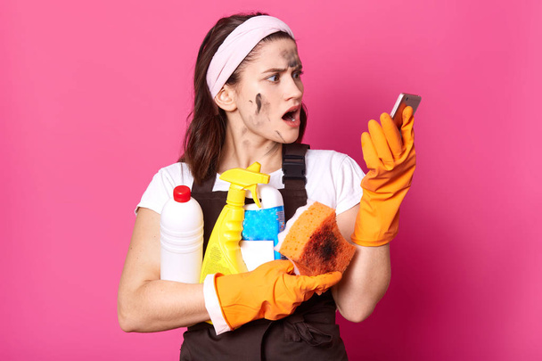 Shocked slender brunette housewife holds bottles of cleaning detergent in one hand and phone in other one, looks shocked about latest news, has unpleasant conversation. Household chores concept. - 写真・画像