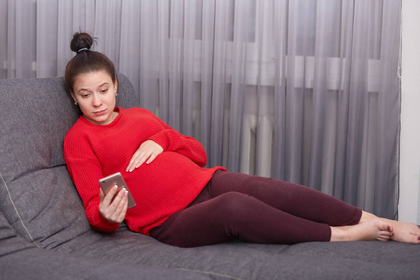 Attentive serious pregnant young lady having rest on sofa, holding one hand on tummy, having phone in her hand, watching video on net, spends time on maternity leave, looks calm and thoughtful. - 写真・画像