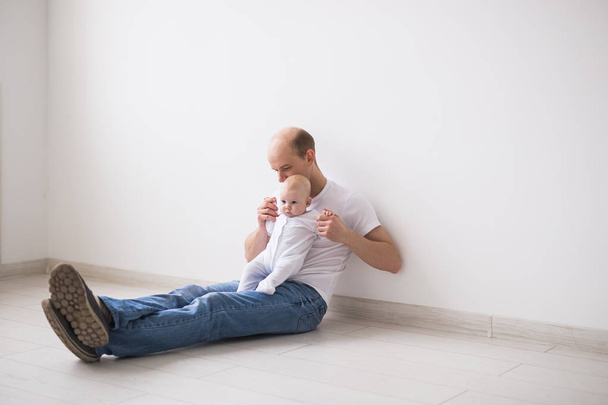 Säugling, Familie und Vaterschaftskonzept - Vater mit Glatze sitzt auf dem Boden und hält sein kleines Baby im weißen Kostüm - Foto, Bild