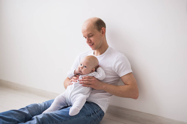 Concept d'enfant, de famille et de paternité - Père chauve assis sur le sol et tenant son petit bébé vêtu d'un costume blanc
 - Photo, image