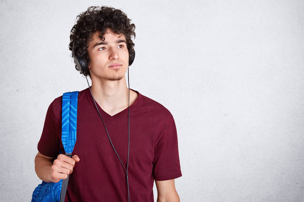 Pensive young guy with headphones on head and blue rucksack, wearing maroon casual t shirt, listens to music, looking thoufhtfully aside, stands isolated over white studio background, Copy space. - Фото, зображення