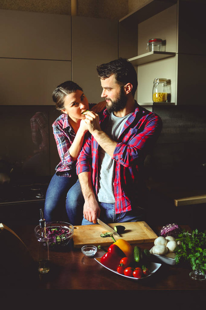 Beautiful young couple in kitchen at home while cooking healthy food. Husband cuts salad and feeds his wife cucumber. Woman looks at the frame. Scene from family life. - Foto, afbeelding