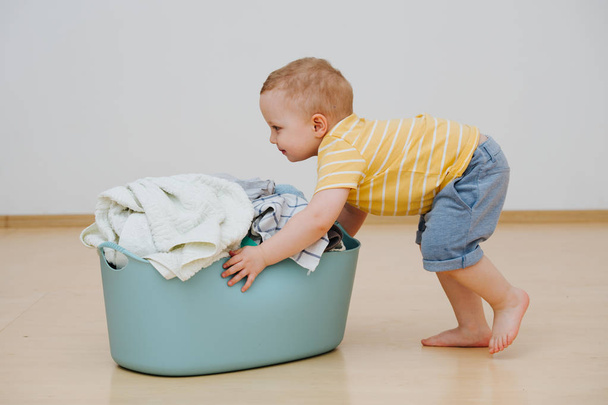 Little toddler boy is moving heavy laundry basin full of washed towels - Foto, Imagem