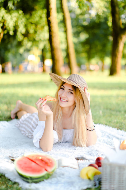 junges lächelndes Mädchen mit Hut und Croissant liegt im Park auf Plaid in der Nähe von Wassermelone. - Foto, Bild