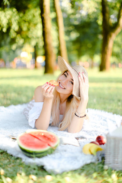 junges glückliches Mädchen mit Hut und Croissant liegt im Park auf Plaid in der Nähe von Watererlemon. - Foto, Bild