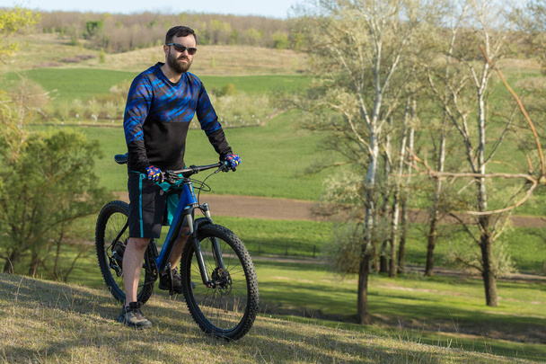 Cyclist in shorts and jersey on a modern carbon hardtail bike with an air suspension fork rides off-road on green hills near the forest - Photo, image