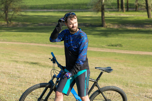 Ciclista en pantalones cortos y jersey en una moderna bicicleta rígida de carbono con una horquilla de suspensión neumática pasea fuera de la carretera en verdes colinas cerca del bosque
 - Foto, Imagen