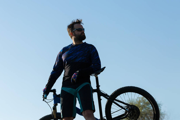 Rowerzysta w krótkich spodenkach i koszulce na nowoczesnym węglowym rowerze z zawieszeniem pneumatycznym z widelcem do jazdy terenowej na zielonych wzgórzach w pobliżu lasu - Zdjęcie, obraz