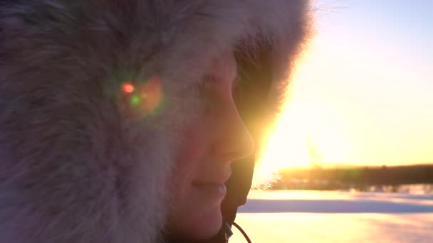 Κοντινό: χαμογελαστή νεαρή γυναίκα με γούνα κουκούλα στο κεφάλι της βλέποντας χρυσό ήλιο ρύθμιση πίσω από το χιόνι τοπίο Λαπωνία. Ευτυχισμένο κορίτσι με ζεστό χειμωνιάτικο μπουφάν την κρύα μέρα την αυγή. Οι άνθρωποι το χειμώνα - Πλάνα, βίντεο