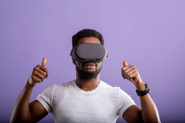 Handsome young African man in VR headset standing against purple background - Foto, afbeelding