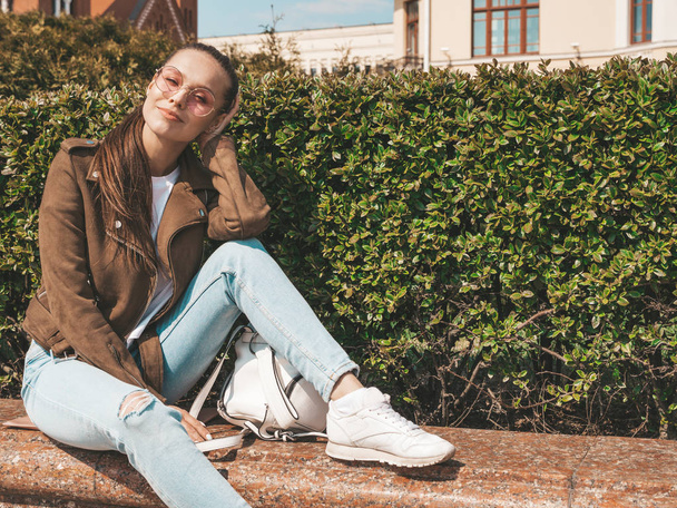 Portrait of beautiful brunette model dressed in summer hipster jacket and jeans clothes. Trendy girl sitting on the bench in the street background. Funny and positive woman in round sunglasses - Foto, immagini