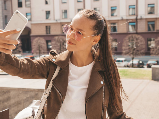 Portrait of beautiful smiling brunette girl in summer hipster jacket. Model taking selfie on smartphone.Woman making photos in warm sunny day in the street in sunglasses - Foto, Bild