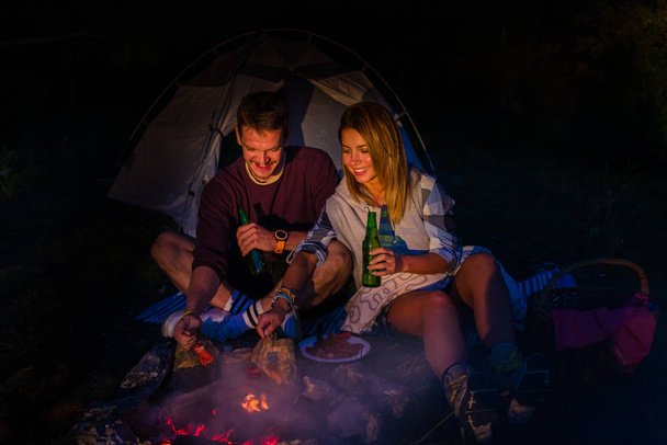 Młoda para piecze kiełbaski na ognisku i pije piwo - Zdjęcie, obraz