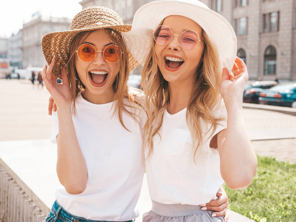 Portrait of two young beautiful blond smiling hipster girls in trendy summer white t-shirt clothes. Sexy carefree women posing on street background. Positive models having fun in sunglasses and hat - Fotoğraf, Görsel