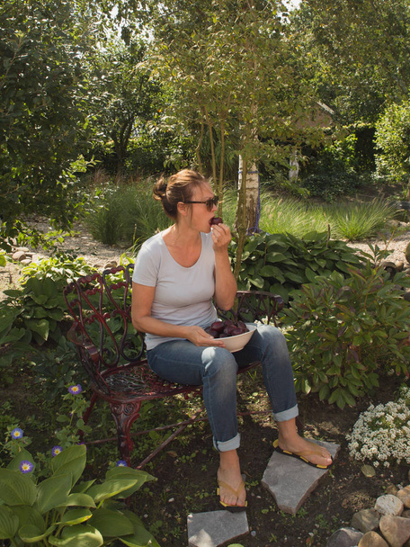 Een mooie vrouw van middelbare leeftijd zit rustig in de tuin en is eten - Foto, afbeelding