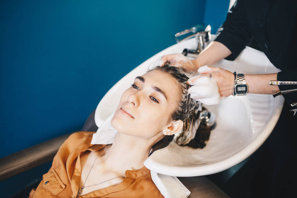 красота, уход за волосами и концепция людей - счастливая молодая женщина с парикмахерской моет голову в парикмахерской
 - Фото, изображение