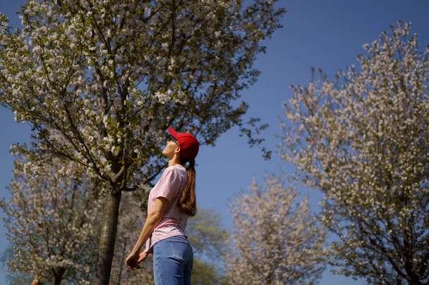 Successful business woman enjoys her leisure free time in a park with blossoming sakura cherry trees wearing jeans, pink t-shirt and a fashion red cap - Photo, Image