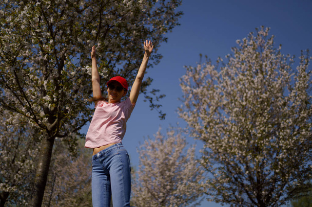 Une femme d'affaires prospère profite de ses loisirs dans un parc avec des cerisiers sakura en fleurs portant un jean, un t-shirt rose et une casquette rouge à la mode
 - Photo, image