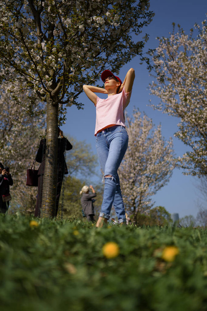 Mulher de negócios bem sucedida desfruta de seu tempo livre de lazer em um parque com árvores florescentes sakura cereja vestindo jeans, camiseta rosa e um boné vermelho moda
 - Foto, Imagem