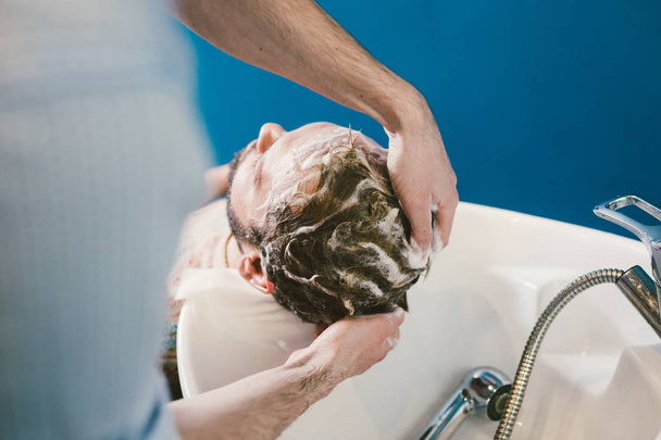 Thema Friseursalon. Friseur zum Haarewaschen in einem Friseursalon. Hände männlich Friseur Nahaufnahme wäscht Haare mit Schaum und Shampoo kaukasischen Mann mit Bart - Foto, Bild