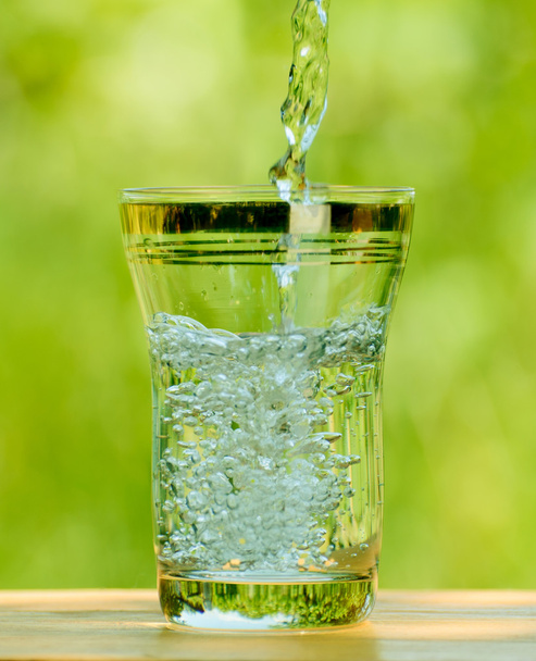 gieten van water in een glas tegen de groene achtergrond - Foto, afbeelding