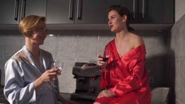 Two women chatting in the kitchen and drinking red wine from glass - One wearing blue morning gown, the other red robe dress - Laughing and smiling - Záběry, video