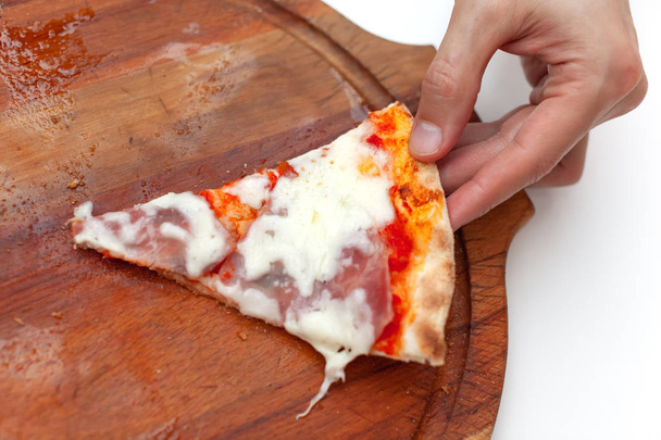 Man took last one slice of delicious Italian pizza. In frame hand taking slice of hot pizza with ham on wooden board on white table. Pizza time. Side view. Close-up. - Foto, immagini