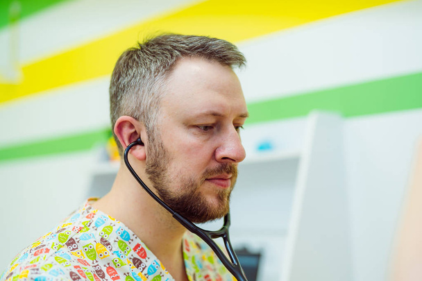 Side view of a male pediatrician with stethoscope in his ears listening to the lungs. Experienced doctor man is checking breath by stethoscope in clinic. Close-up - Foto, afbeelding