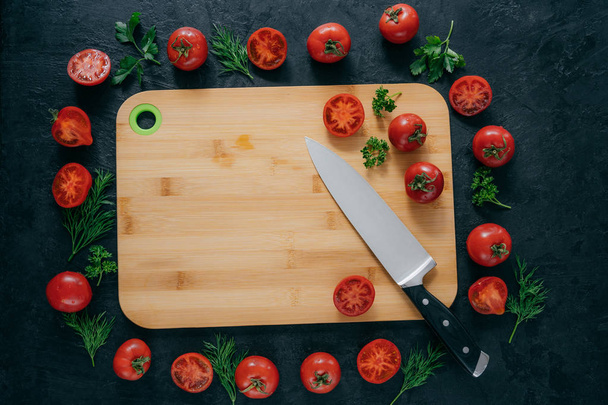 Ντομάτες κορνίζα γύρω από ξύλινη σανίδα κοπής. Ώριμα λαχανικά και φέτες, πράσινο μαϊντανό και άνηθο κοντά σε σανίδα κουζίνας και μαχαίρι. Τέχνη τροφίμων - Φωτογραφία, εικόνα