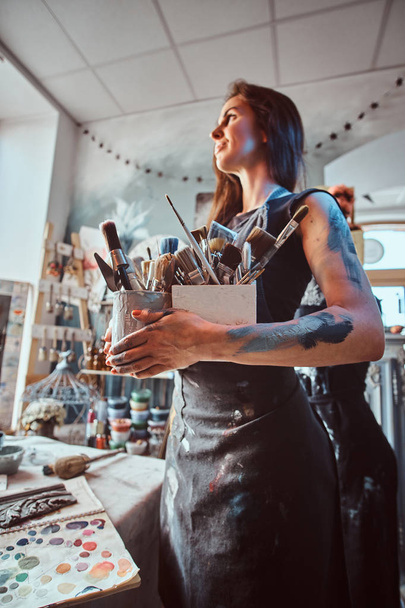 junge attraktive Malerin posiert mit Pinseln in ihrer eigenen gemütlichen Werkstatt - Foto, Bild