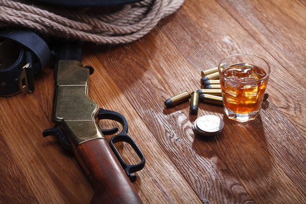 Wild west rifle and ammunitions with glass of whisky and ice with old silver dollar on wooden table - Фото, изображение