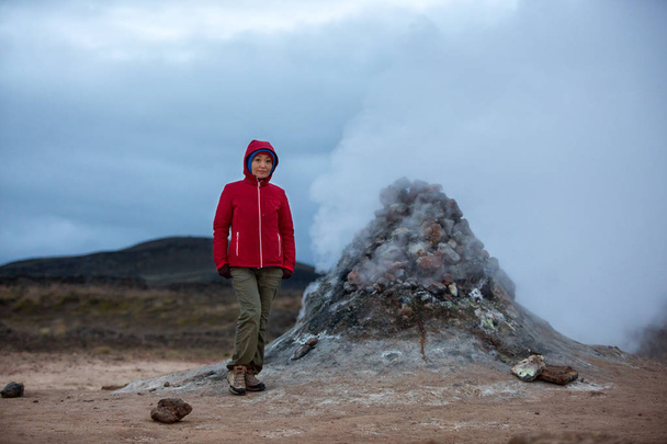A girl tourist in a red jacket stands against the background of sulfuric smoke erupting from the ground. - Фото, зображення