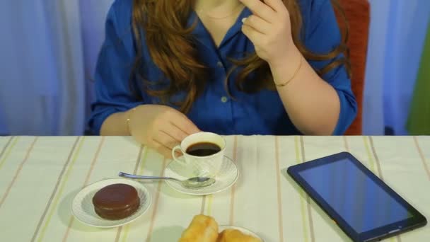 woman in a cafe at a table drinking coffee with dessert and reading the news on the tablet - Imágenes, Vídeo