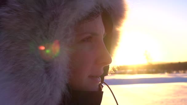 Zbliżenie: uśmiechnięta młoda kobieta z futerkiem na głowie oglądając złote słońce ustawienie za Snowy krajobraz Laponii. Happy Girl w ciepłej zimowej kurtki w zimny dzień na poranny wschód słońca. Ludzie w zimie - Materiał filmowy, wideo