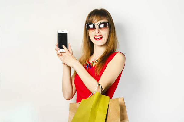 Mladá dívka v červených šatech s telefonem a nákupními taškami na světlém pozadí. Online nákupní koncepce, nákupní aplikace, prodej. - Fotografie, Obrázek