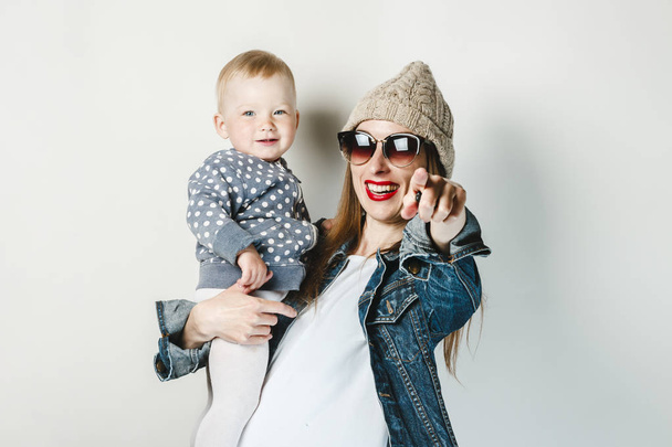 A young girl in a denim jacket, glasses and a hat holds a small child on a white background. Concept game with children, happy family, joy and fun, young family. - Фото, зображення