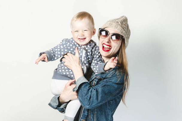 A young girl in a denim jacket, glasses and a hat holds a small child on a white background. Concept game with children, happy family, joy and fun, young family. - Photo, Image