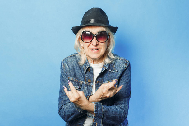 Старушка в джинсовой куртке, солнцезащитных очках и шляпе делает жест на скале на синем фоне. Концепция модная бабушка, старуха, экшн рокеров Коза
. - Фото, изображение