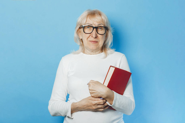 Una anciana en gafas con una cara feliz sosteniendo un libro en sus manos sobre un fondo azul. Concepto anciana lee libros, educación, club de lectura
. - Foto, imagen