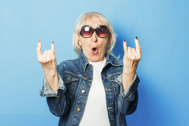 Kot ceketli yaşlı bir kadın, güneş gözlüğü mavi bir arka plan üzerinde kaya üzerinde bir jest yapar. Konsept moda büyükanne, yaşlı kadın, aksiyon rockçılar Keçi. - Fotoğraf, Görsel