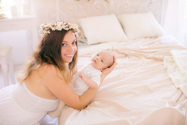 Young cute mom in a wreath of cherry blossoms in a white dress holding a baby in her arms in a spring bedroom with blossoming cherry branches - Foto, Bild