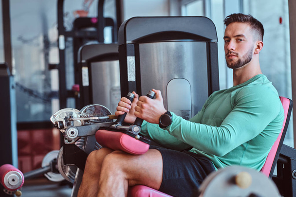 Forte homem pensativo está sentado no aparelho de treinamento no ginásio e fazendo exercícios de pernas
 - Foto, Imagem