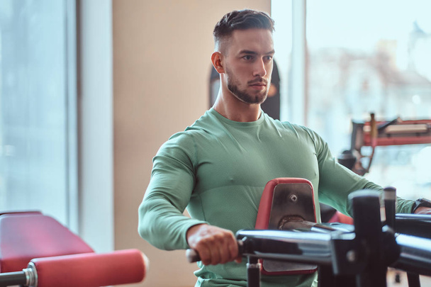 Young pensive bodybuilder in green shirt is doing exercises on training apparatus - Φωτογραφία, εικόνα