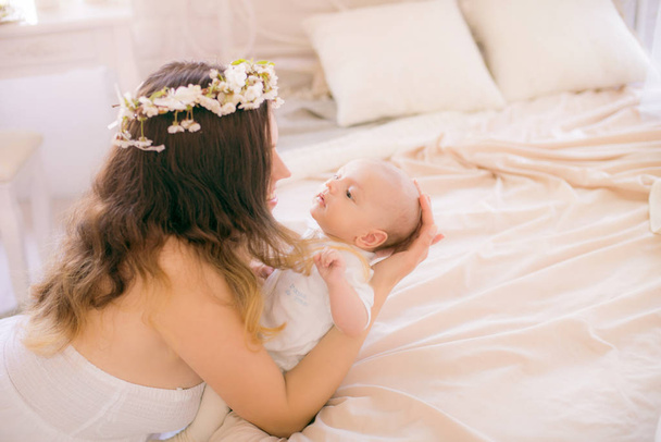 Young cute mom in a wreath of cherry blossoms in a white dress holding a baby in her arms in a spring bedroom with blossoming cherry branches - Fotó, kép