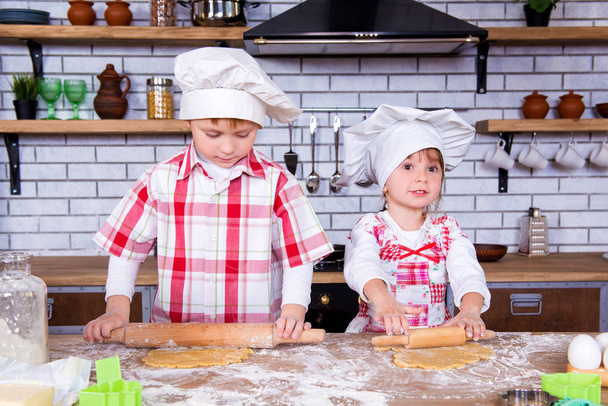 Children brother and sister bake gingerbread in the kitchen. A girl and a boy roll out the dough with a rolling pin on the table - Fotoğraf, Görsel
